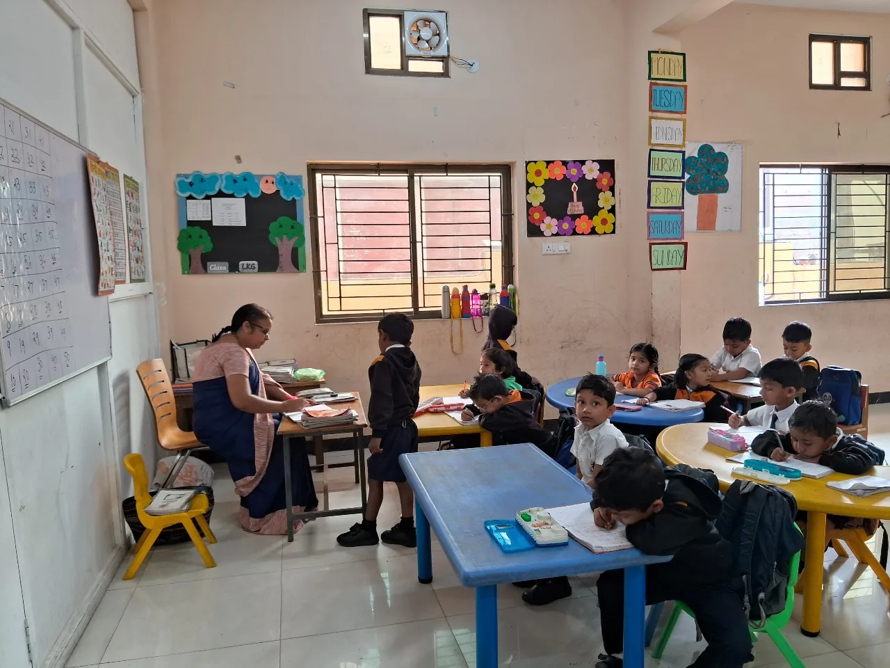A kindergarten (LKG) classroom at Cardinal Warriors with a teacher and young children at bright activity tables, walls decorated with days-of-the-week and learning charts.