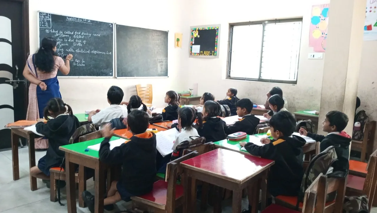A primary (Class I) classroom at Cardinal Warriors: a teacher writing on the blackboard while young cadets in uniform sit at colourful desks.