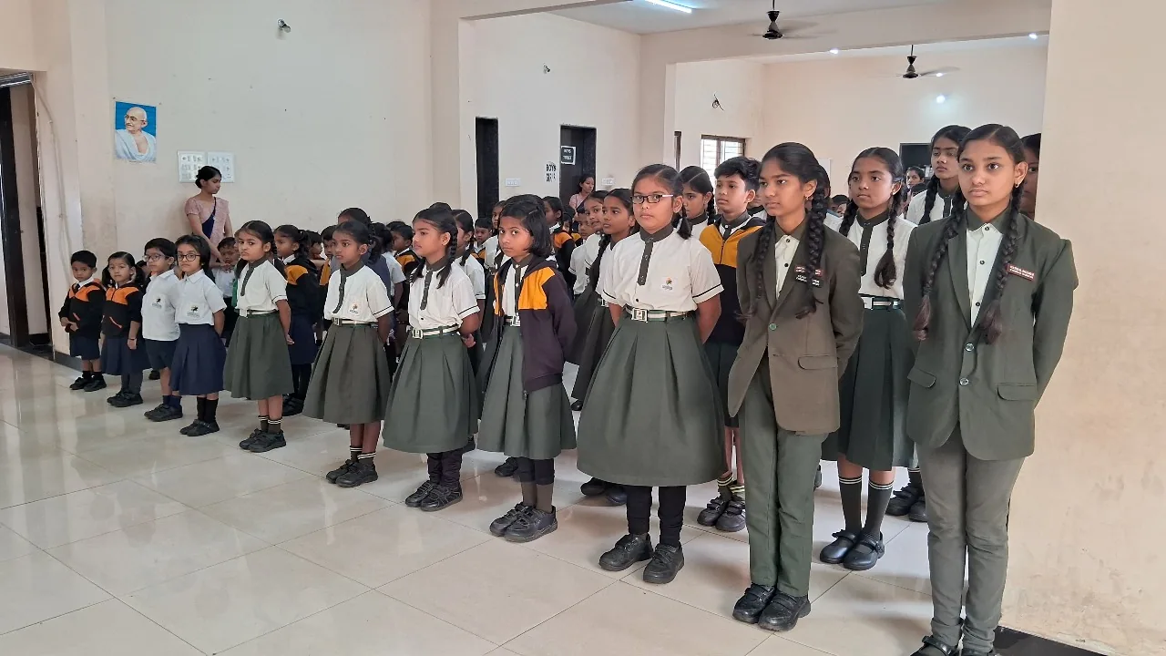 Students of all ages in Cardinal Warriors uniform standing in disciplined rows for an indoor morning assembly, a portrait of Mahatma Gandhi on the wall.