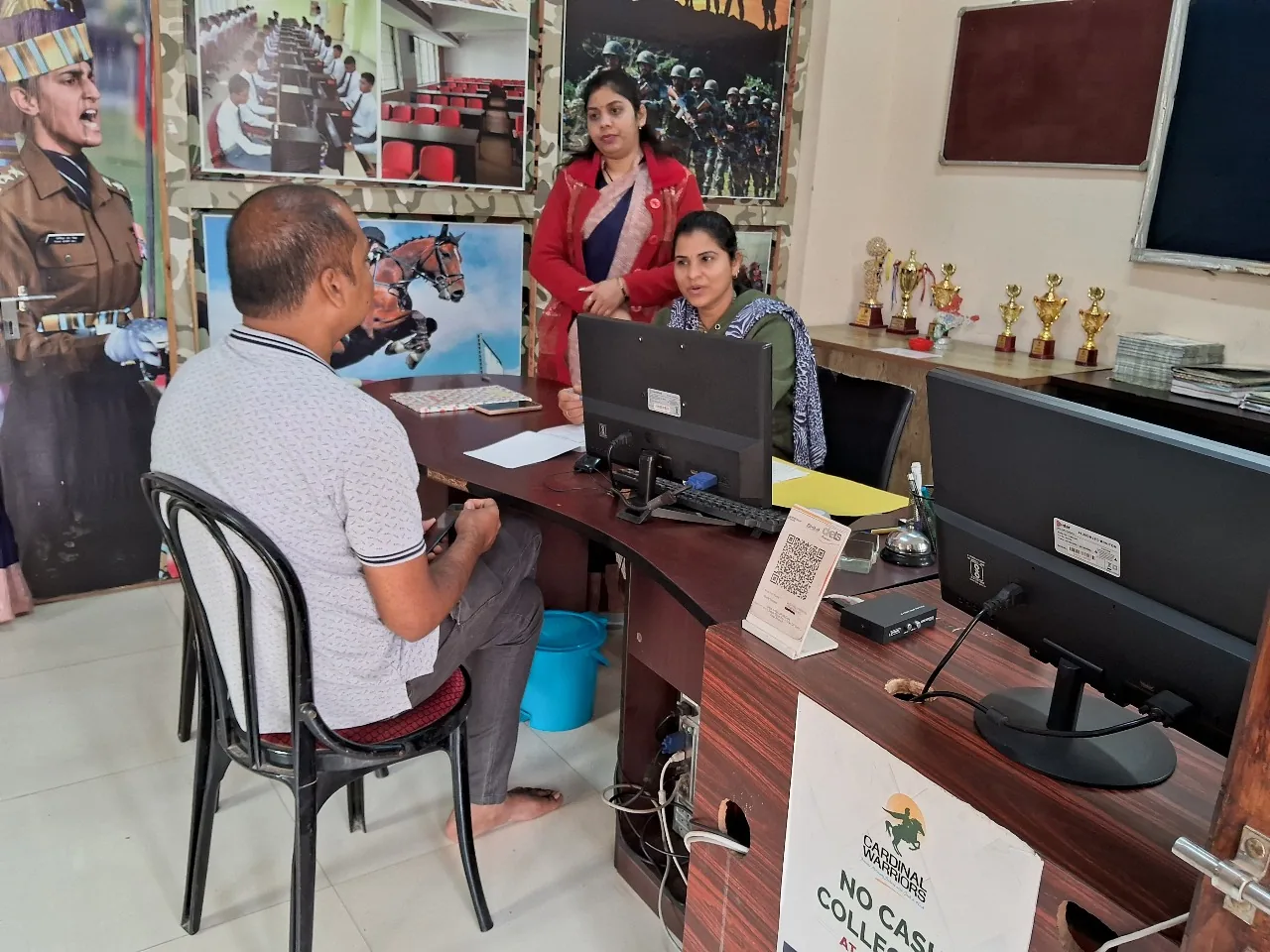 A parent meeting admissions staff at the Cardinal Warriors front desk, with cadet posters, trophies and an online-fees notice in the background.