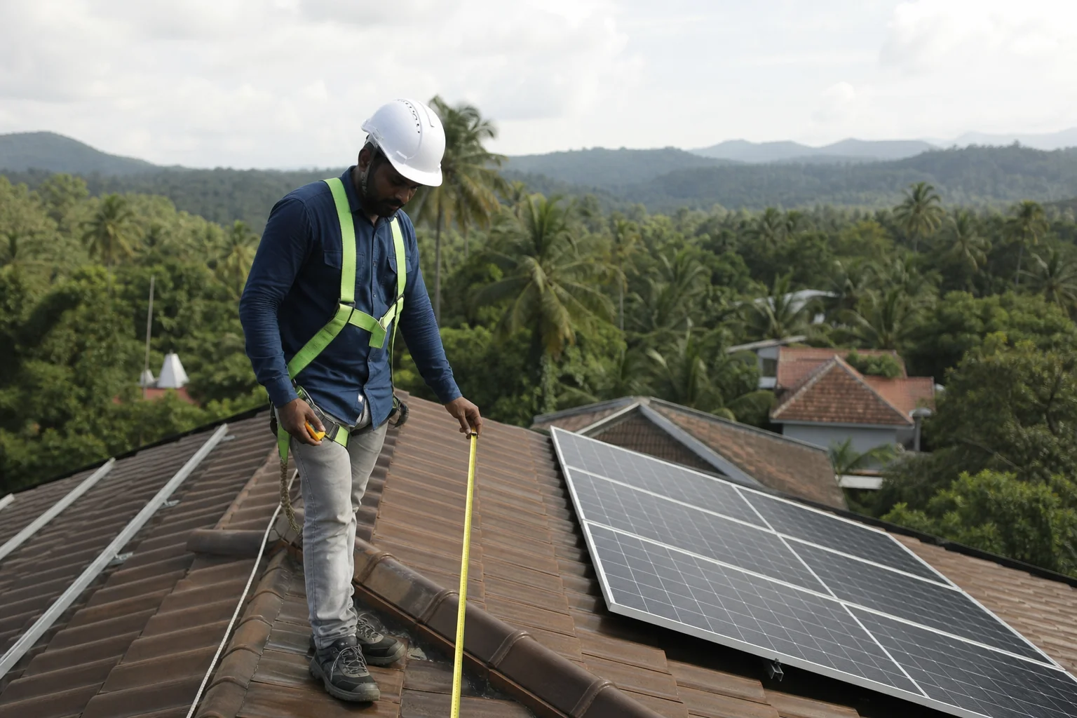 Technician measuring or inspecting rooftop space for solar installation in a residential Kerala setting. Photorealistic photo of a solar technician in work clothes and safety gear standing on a residential rooftop in Kerala, measuring roof