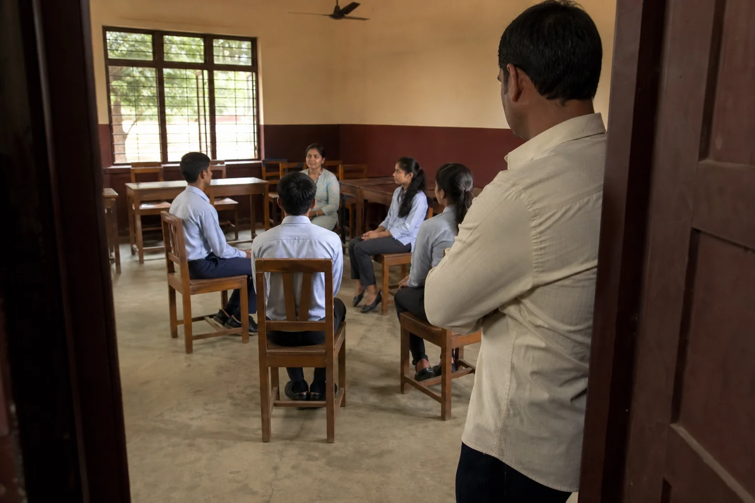 photorealistic photo of an Indian parent in casual formal clothing standing at the doorway of a classroom in a boarding school in Chhattisgarh, watching a small group of students practicing a mock interview seated in a semi-circle with a