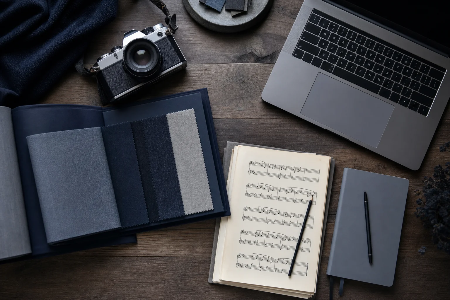 Photorealistic photo of an organized creative studio desk from overhead, featuring a camera, a fabric swatch book, a musical notation sheet, and a laptop, arranged neatly on a wooden surface, soft natural light, cool professional color