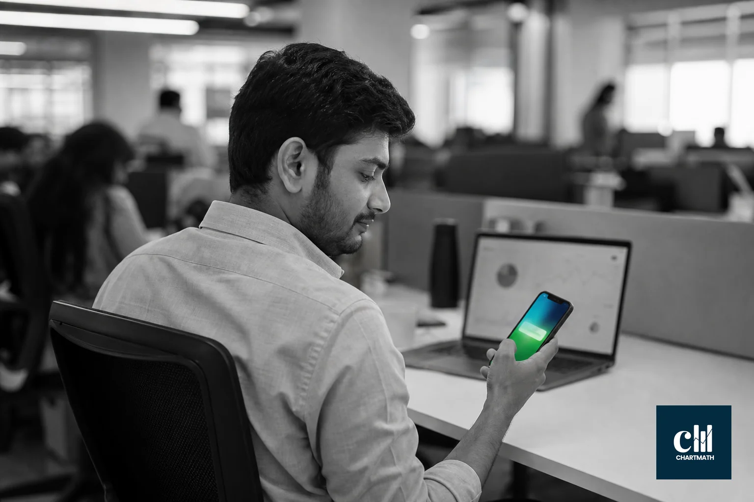 photorealistic photo of a young Indian professional in business casual clothing sitting at a desk in a modern open-plan office in India, glancing calmly at a smartphone showing a notification, laptop with charts blurred in the background