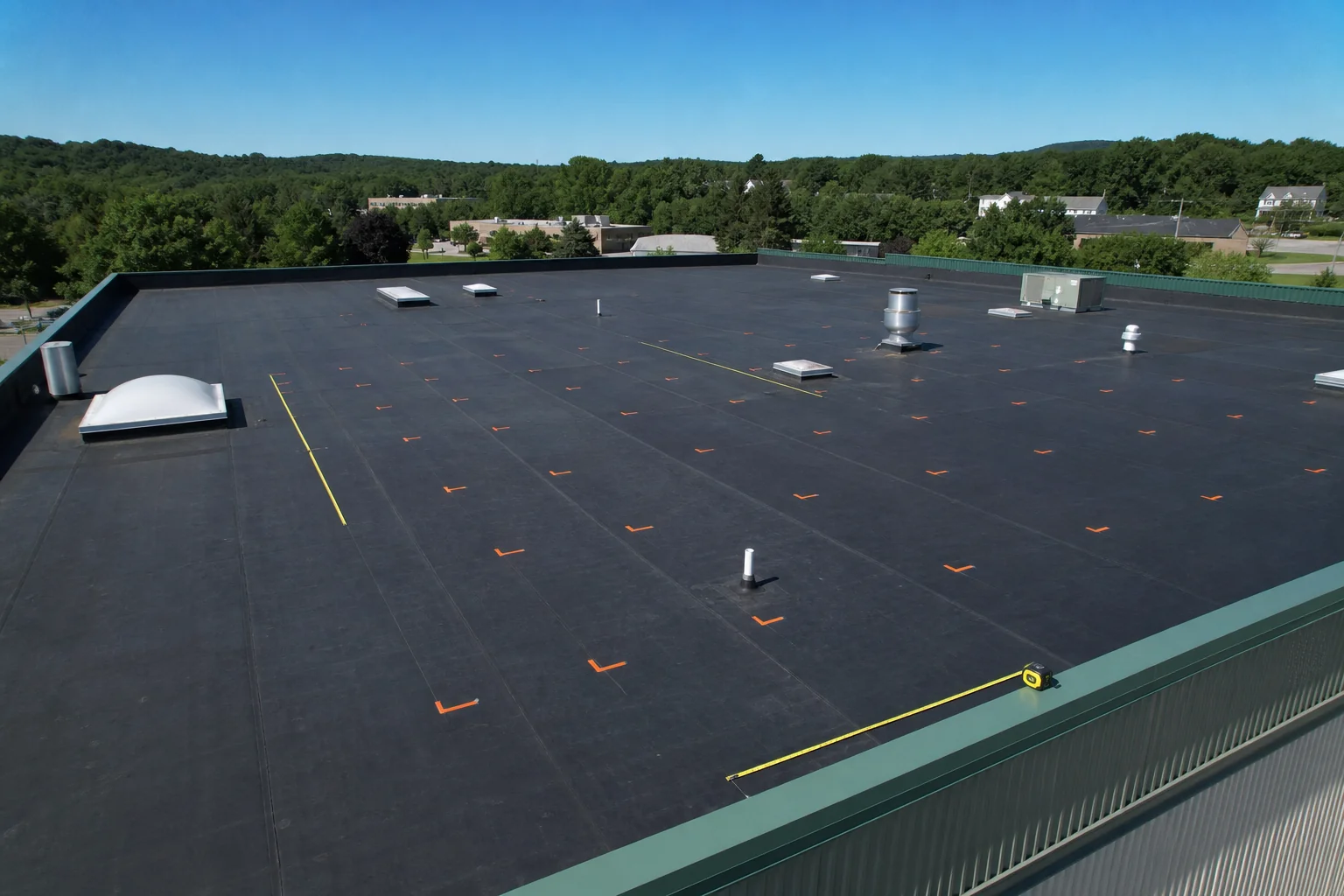 Aerial view of a flat commercial rooftop being measured for solar panel layout. Photorealistic aerial drone photo of a flat commercial warehouse rooftop in New England with a measuring tape and layout markers laid out, clear blue sky, dark