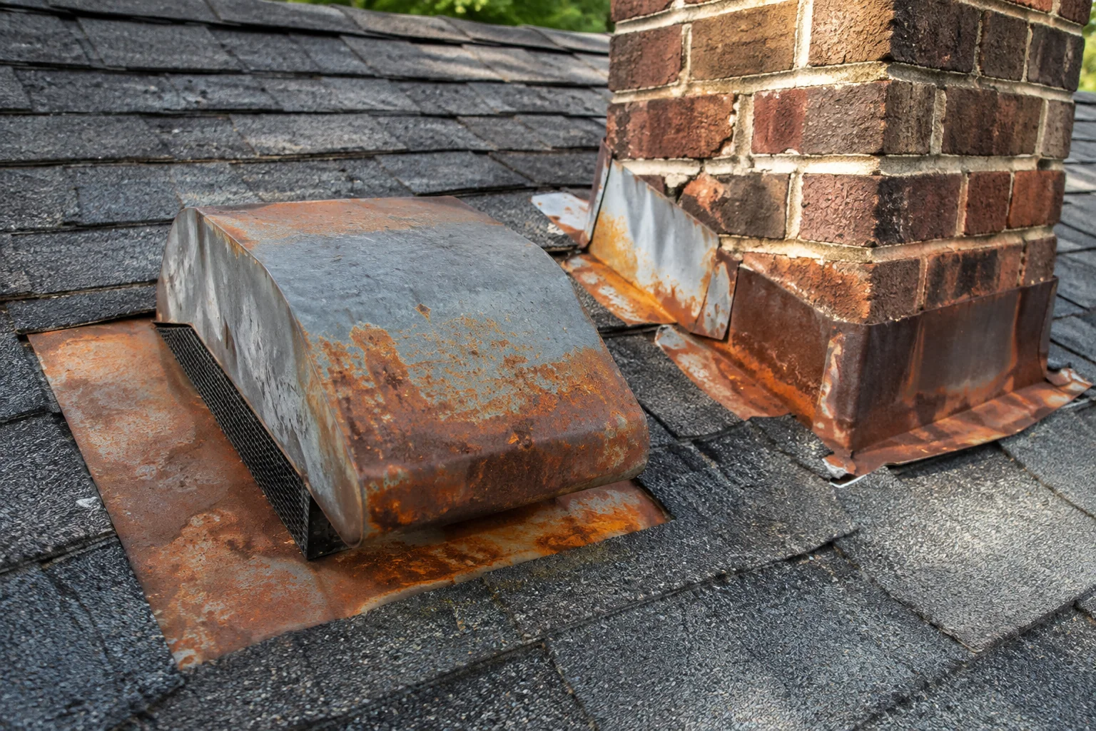 Close-up of corroded metal roof flashing around a chimney with worn shingles