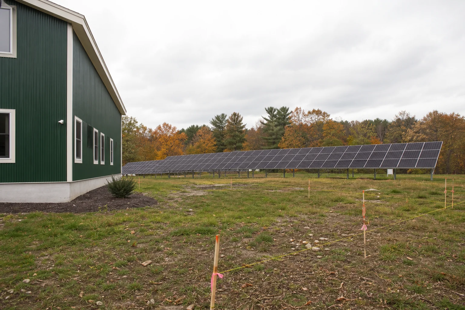 Commercial ground mount solar array on New England business property with zoning survey stakes. photorealistic photo of a commercial ground-mounted solar panel array installed on open land behind a small New England business building