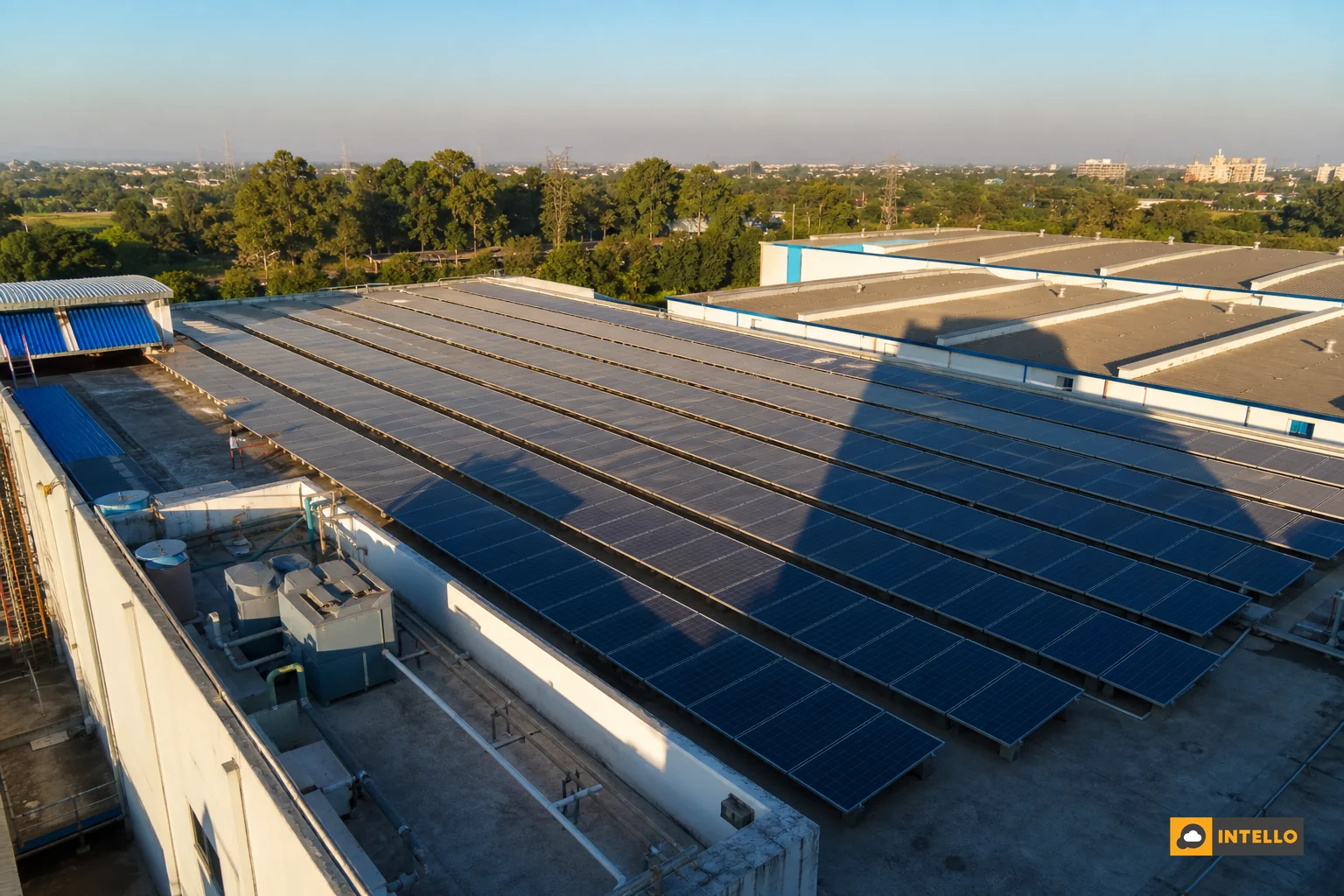 Aerial view of commercial rooftop solar array with tree and building shadows falling across panels. Photorealistic aerial drone photo of a large commercial rooftop solar installation in India, showing long shadows cast by a nearby building