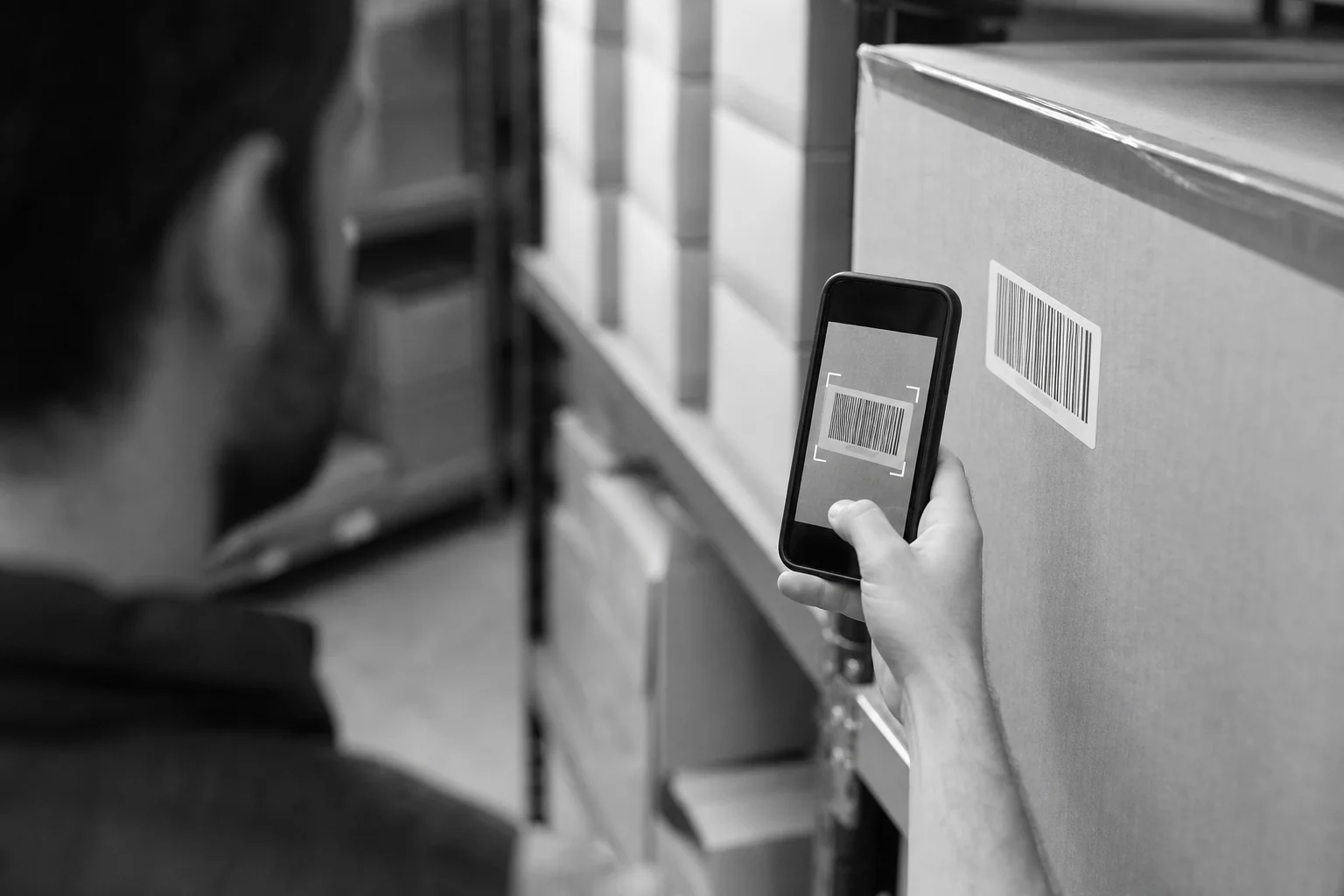 Warehouse worker scanning barcode with phone for stock tracking. Photorealistic photo of a warehouse staff member scanning a product barcode using a smartphone camera, boxes and shelving in the background, black and white toned photography