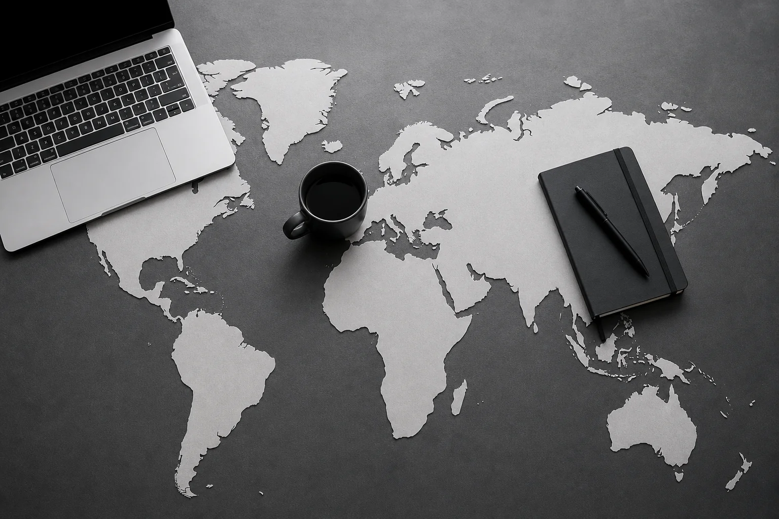 photorealistic: An overhead flat-lay photo of a world map made of paper cutouts with a laptop, a coffee mug, and a notebook placed near the UK and India regions, black and white monochrome palette with minimal contrast lighting, clean