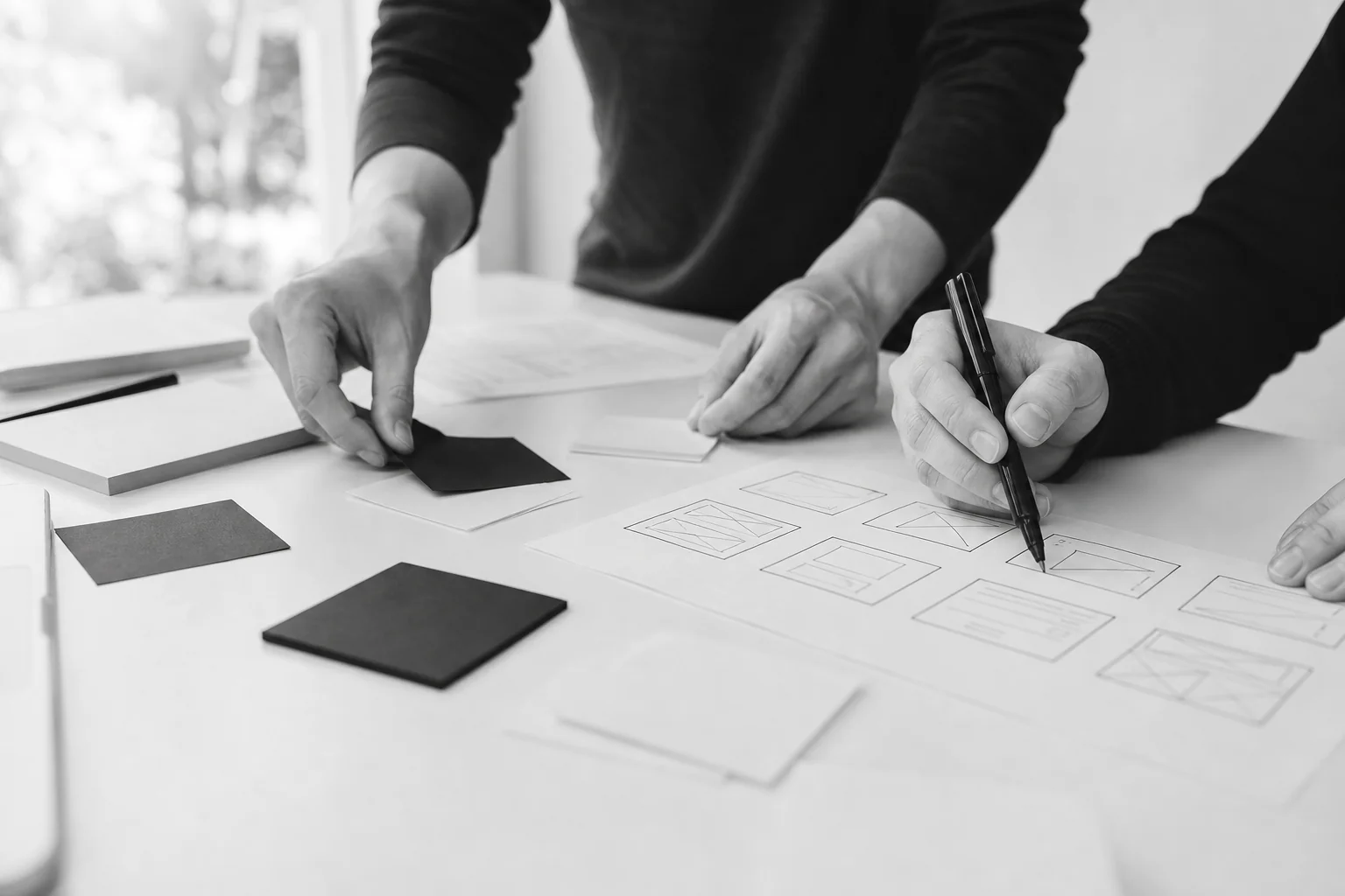 Close-up of hands sketching wireframes and sticky notes during a requirements workshop. Photorealistic photo, close-up shot of hands arranging blank sticky notes and sketching simple wireframe boxes on paper during a planning workshop