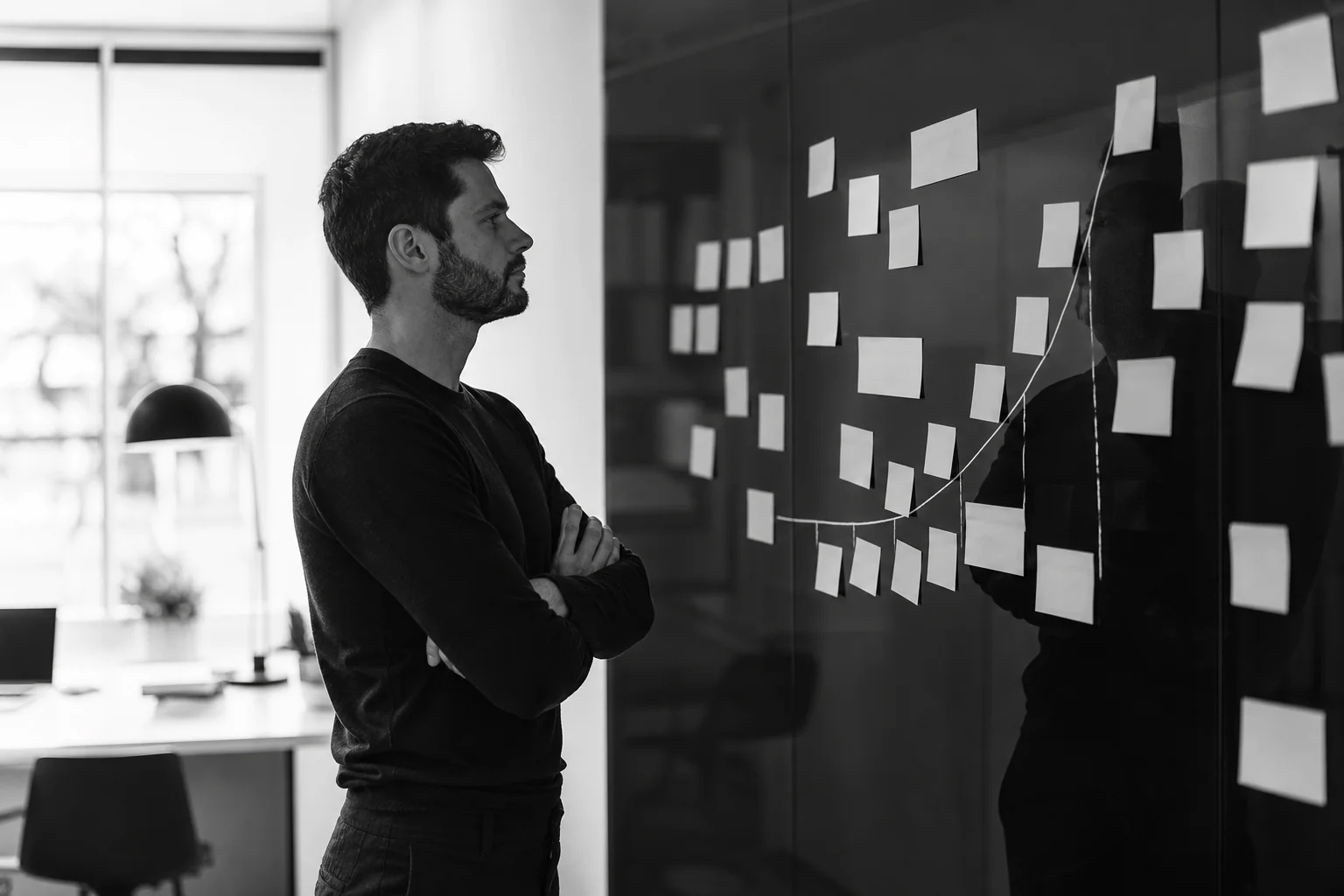 photorealistic: A founder standing at a black glass wall covered in printed sticky notes and a simple hand-drawn growth curve, arms crossed, thoughtful expression, looking at the roadmap, dressed in smart casual clothing, bright minimalist