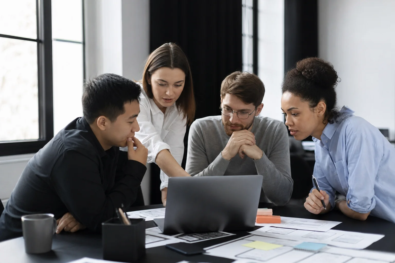Development team collaborating around a laptop reviewing app design wireframes. photorealistic, natural photography style: a small team of developers and designers gathered around a laptop and printed wireframes on a desk, discussing a