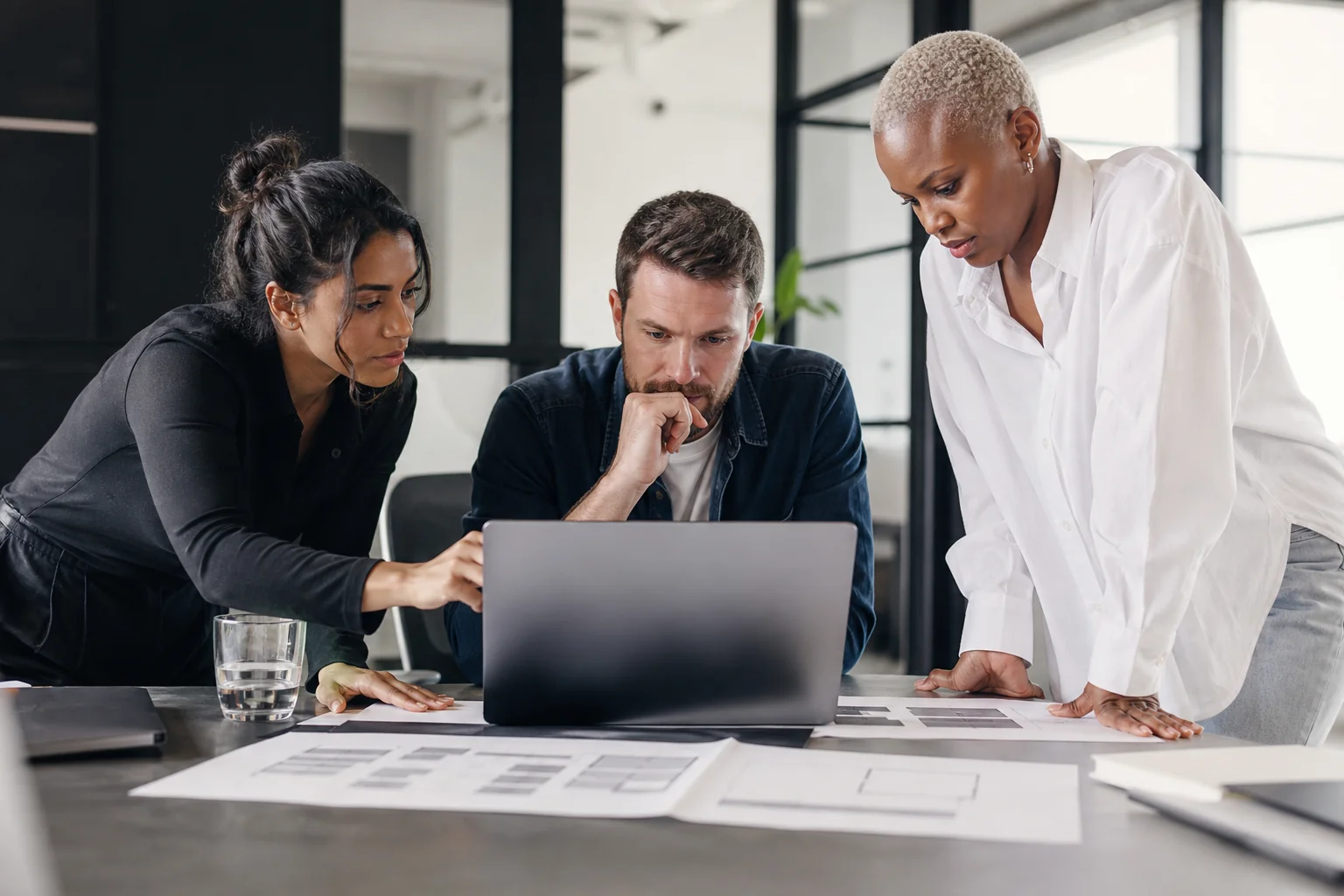 Diverse engineering team collaborating around a laptop reviewing system architecture diagrams. Photorealistic photo: a small diverse software engineering team of three people gathered around a laptop and printed architecture diagrams in a