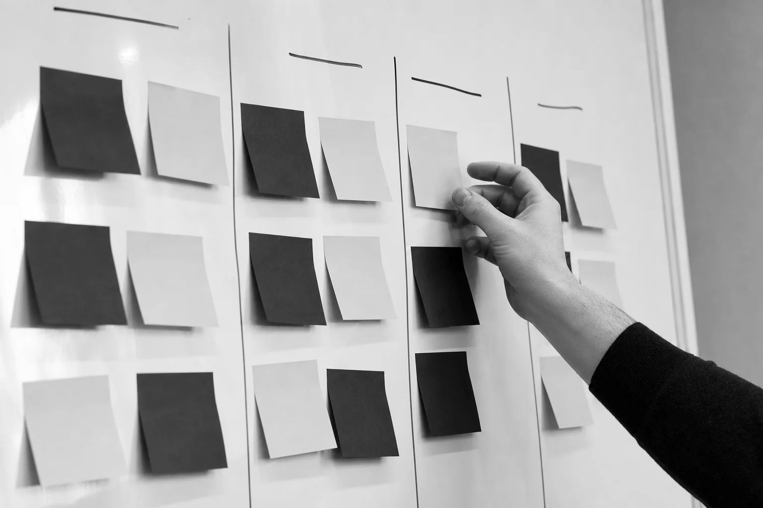 Whiteboard session sorting sticky notes into priority columns. photorealistic photo of a whiteboard covered with black and white sticky notes arranged into four labeled columns, a person's hand placing a note into a column, close-up