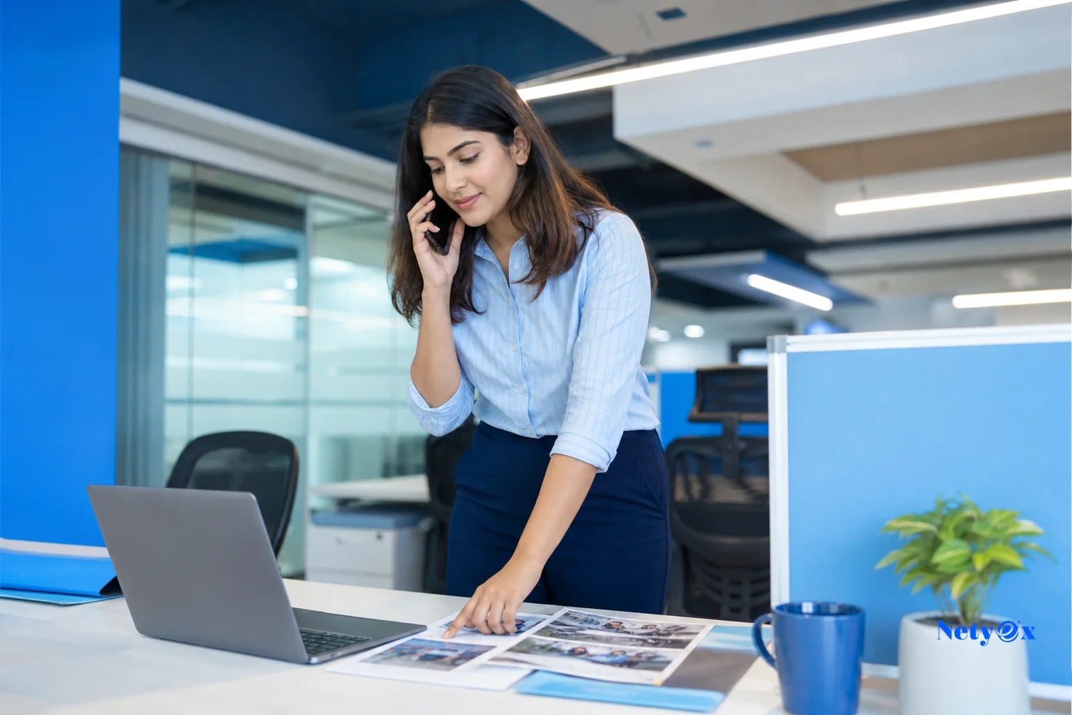 photorealistic photo of a sourcing coordinator in a modern office in Noida India, standing at a desk with a laptop and printed supplier photos spread out, phone in hand as if coordinating logistics, bright modern office with blue accent