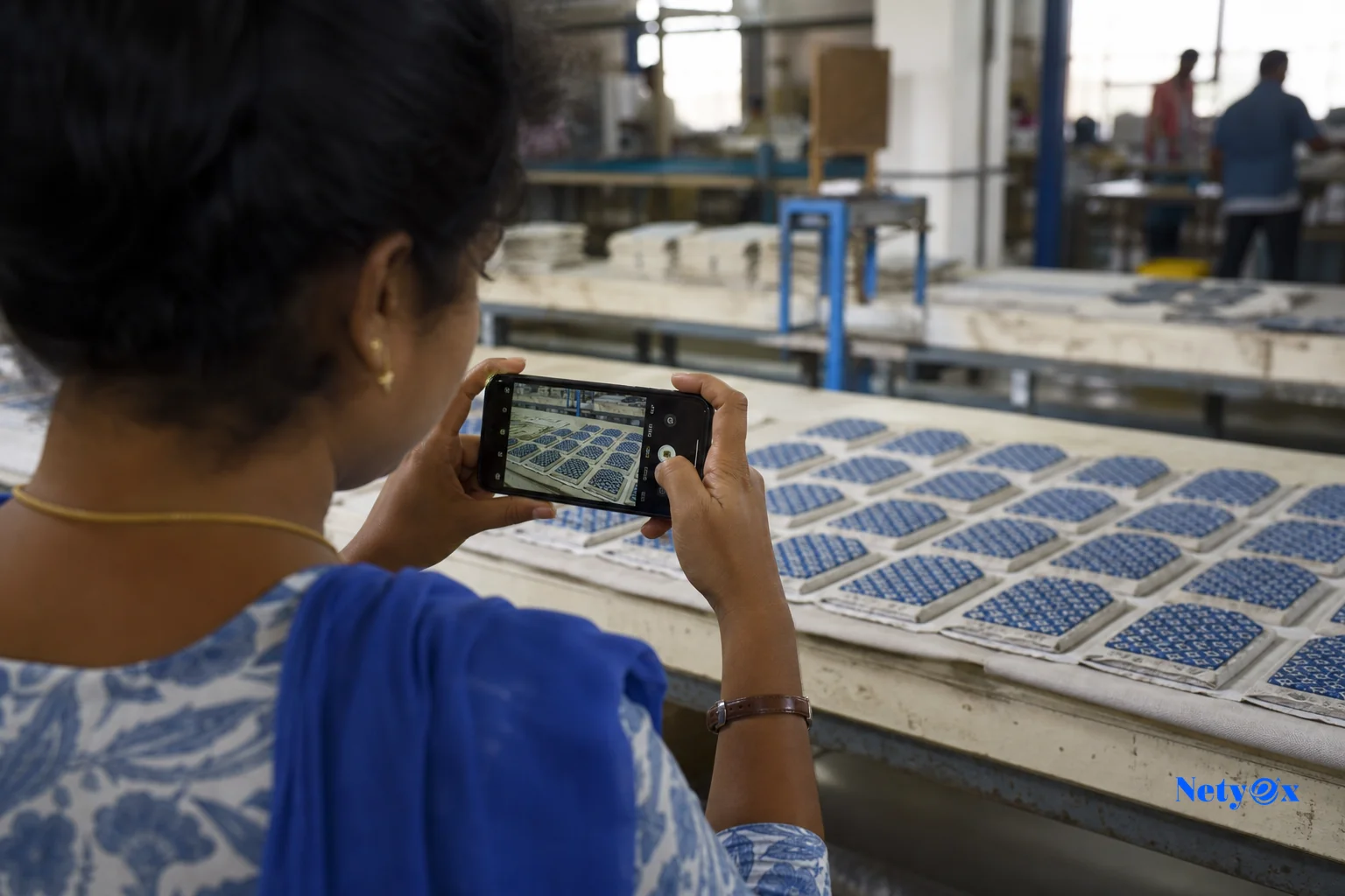 photorealistic: Close-up view over the shoulder of a factory quality staff member in India photographing rows of partially finished handicraft or textile products on a production line using a smartphone, industrial workshop background with