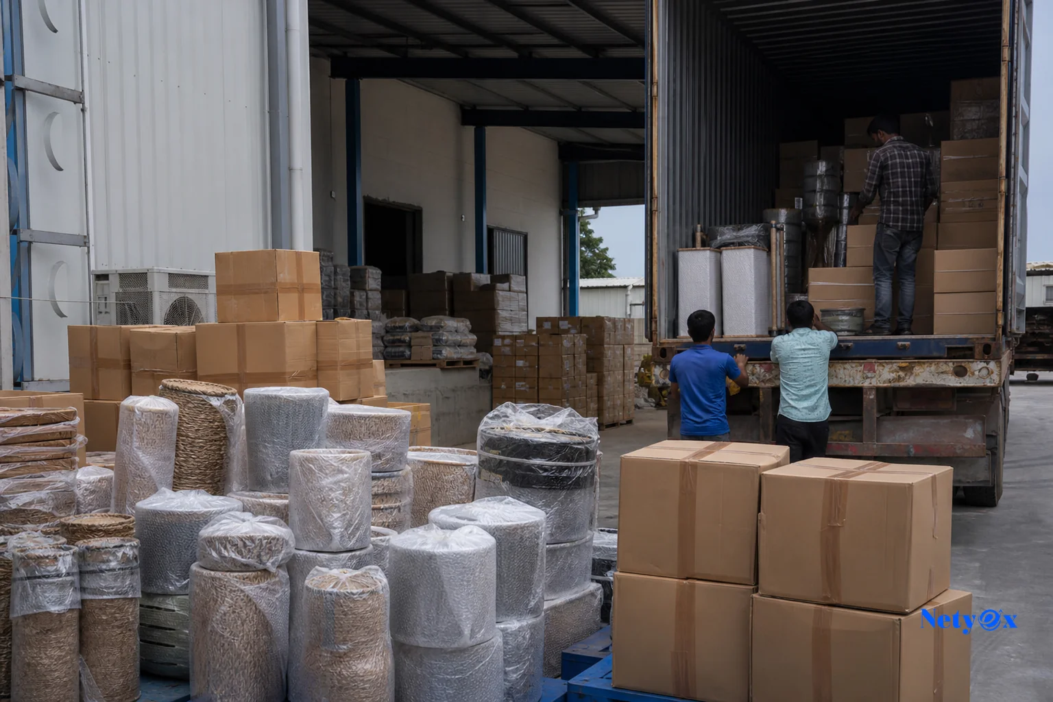 photorealistic photo of stacked export cartons and pallets of home decor products being loaded onto a truck at an Indian logistics warehouse, workers in the background, natural daylight, color palette leaning toward slate blue and grey
